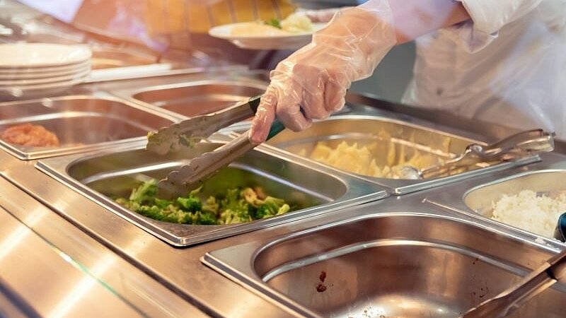 Eine Person schöpft Essen aus Warmhaltebehältern an einem Buffet.