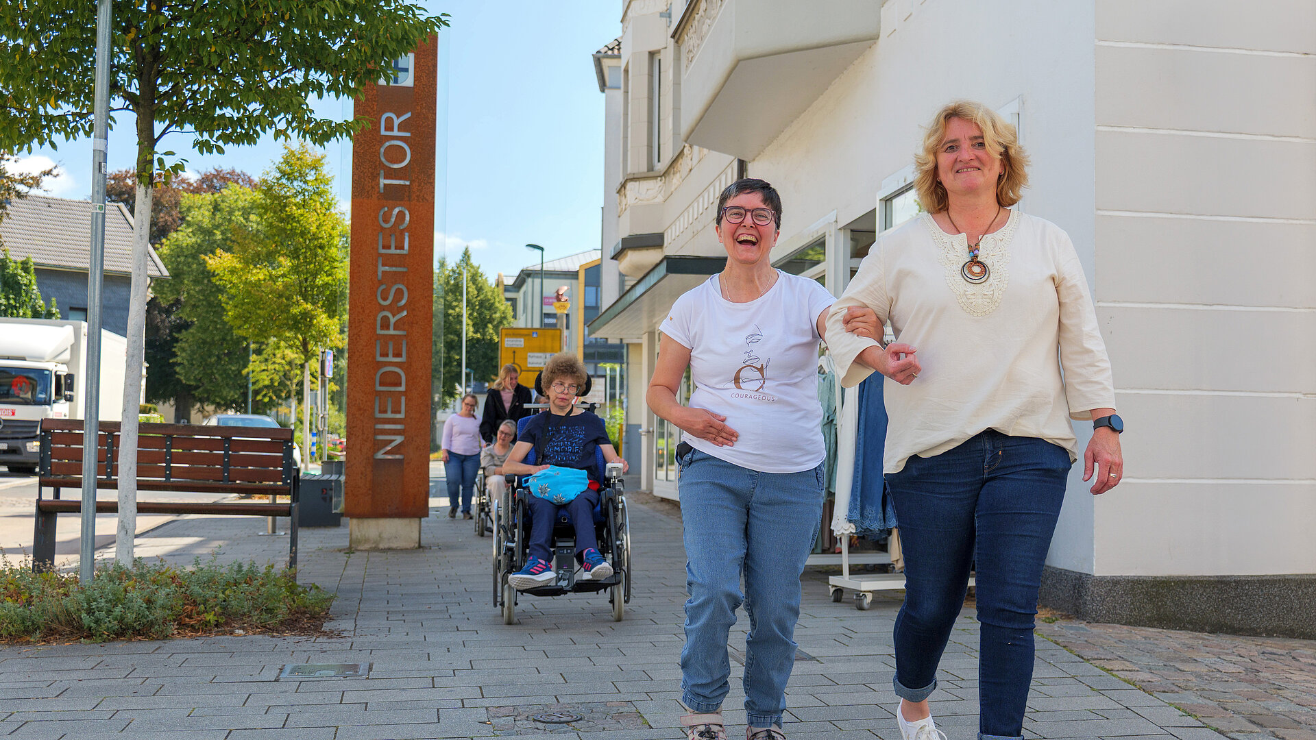 Zwei Frauen gehen untergehakt und lachend durch die Fußgängerzone, dahinter eine Frau im Rollstuhl mit Begleitung.