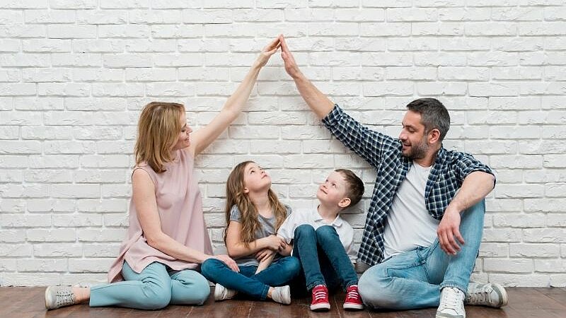 Eine vierköpfige Familie sitzt gemeinsam auf dem Boden. Die Eltern und ihre beiden Kinder strecken spielerisch die Arme nach oben.