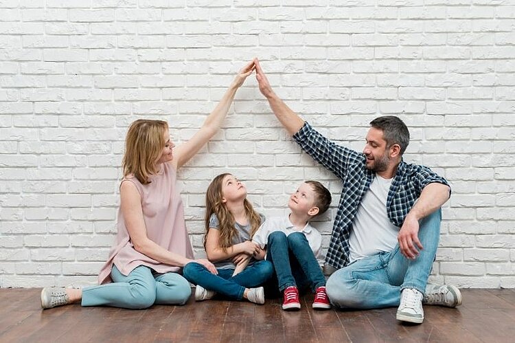 Eine vierköpfige Familie sitzt gemeinsam auf dem Boden. Die Eltern und ihre beiden Kinder strecken spielerisch die Arme nach oben.