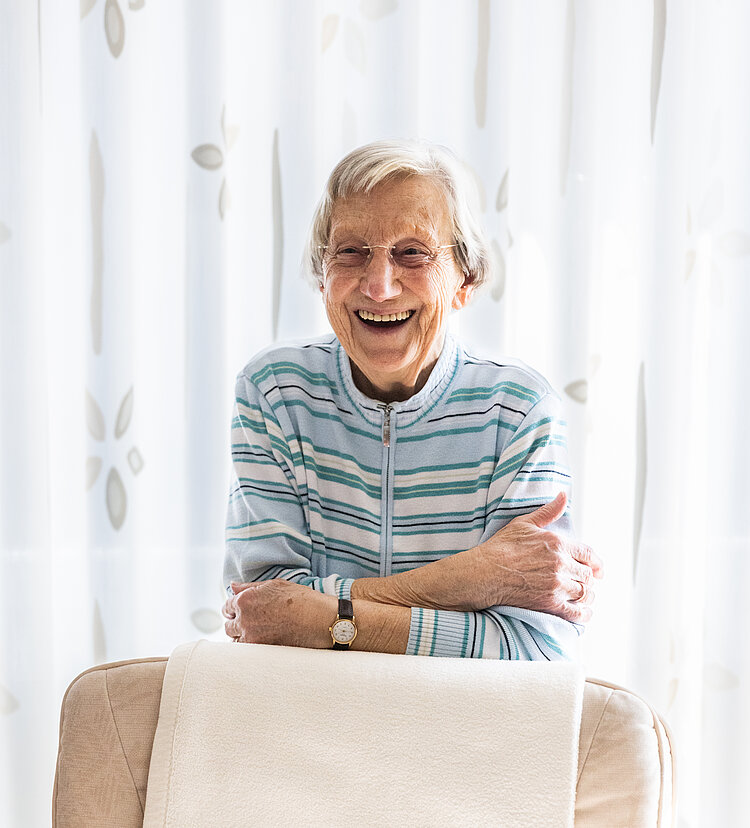 Seniorin stützt sich lachend von hinten auf ihren Sessel