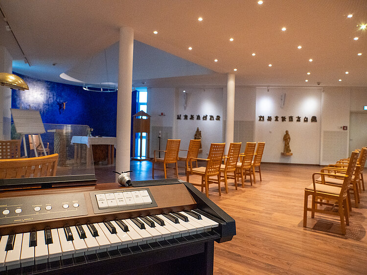 Blick in die bestuhlte Kapelle, mit der Orgel im Vordergrund. An den weißen Wänden hängen kleine Skulpturen