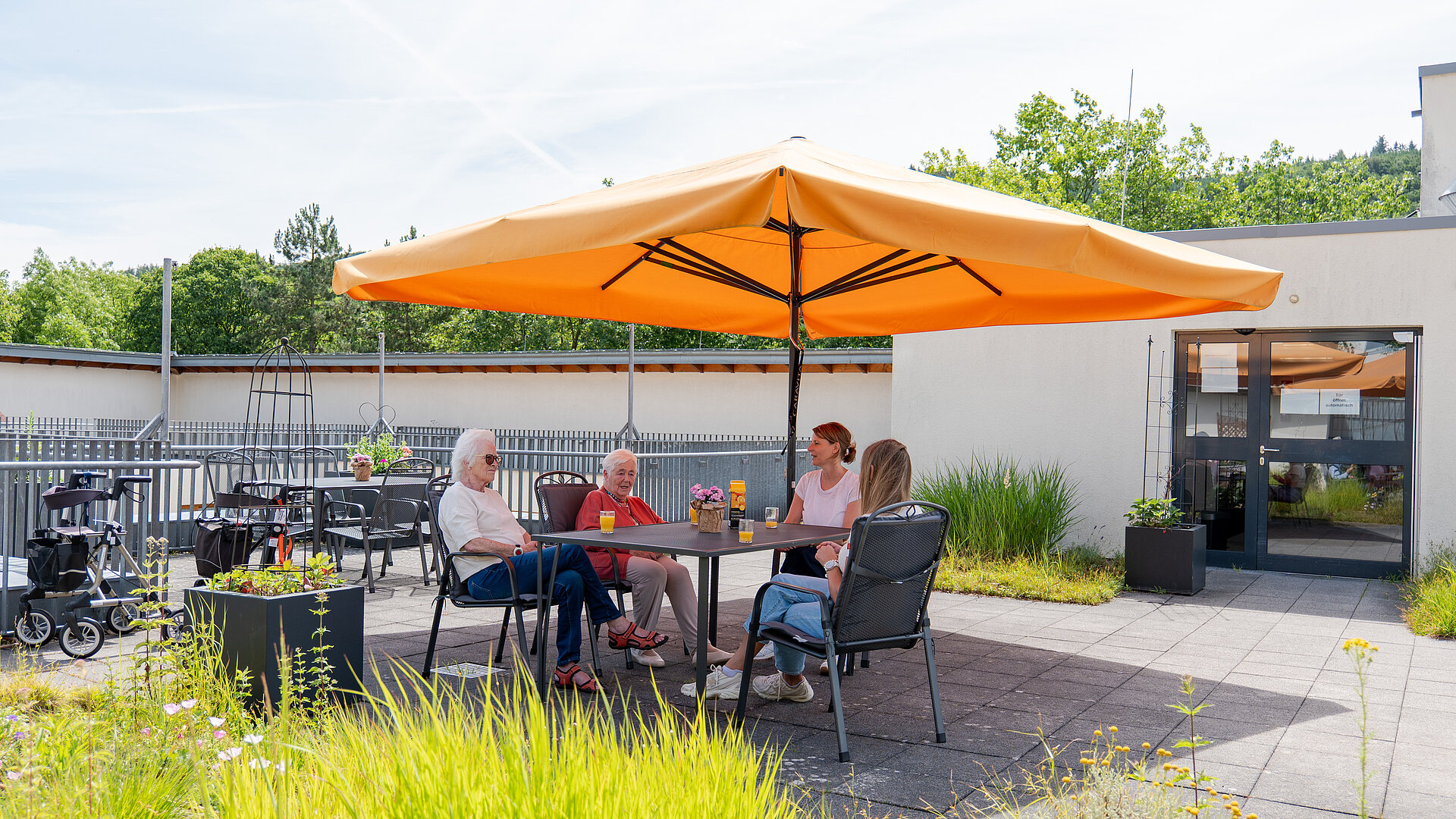 Vier Personen sitzen an einem Tisch unter einem großen orangefarbenen Sonnenschirm auf einer Terrasse mit Pflanzen und Blick auf ein modernes Gebäude