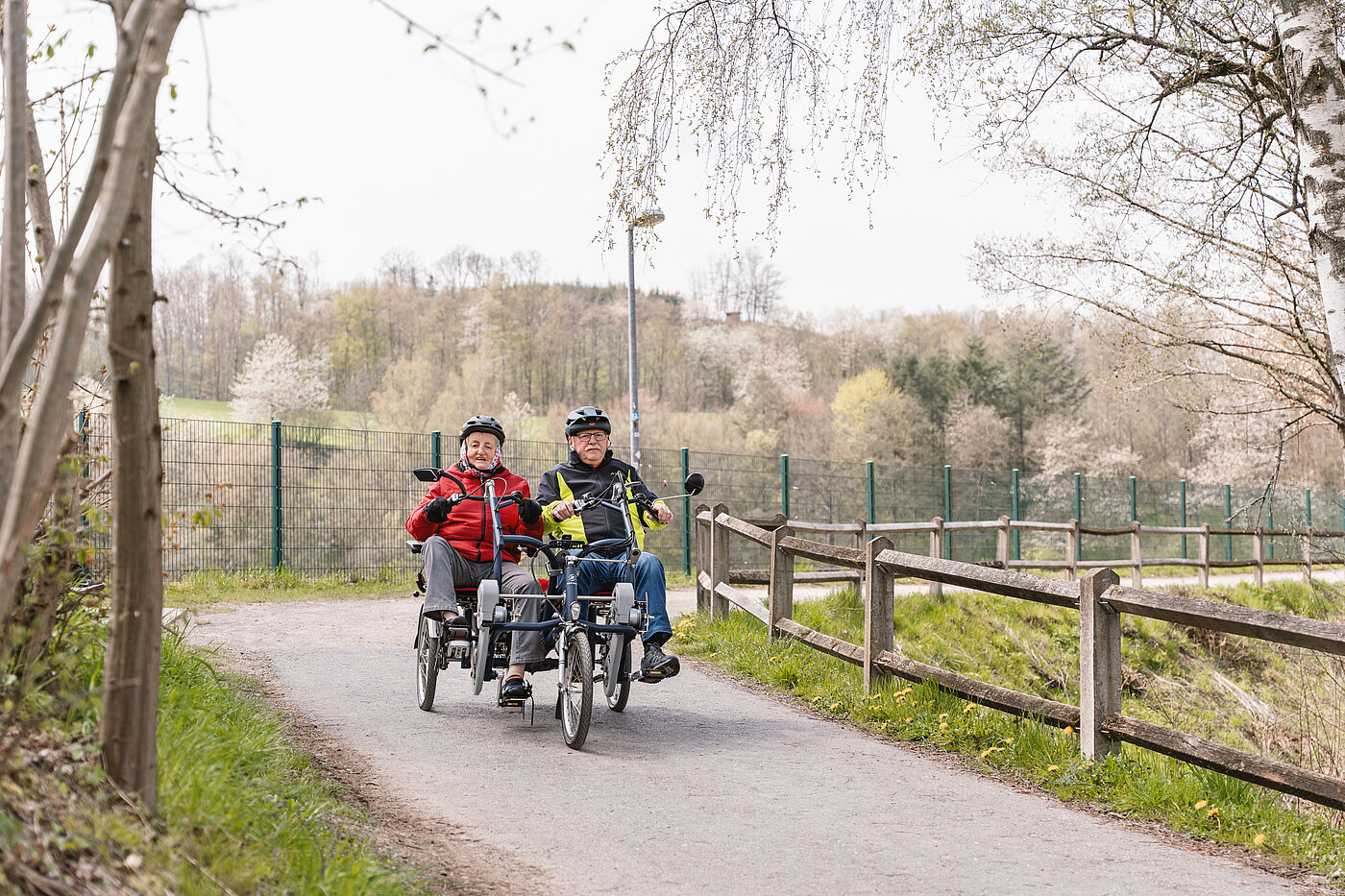 Zwei ältere Personen fahren auf einem Tandemfahrrad auf einem Weg umgeben von einem Holzzaun und Bäumen