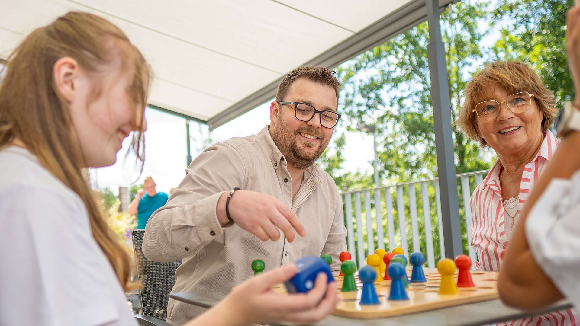 Ein Mann, eine junge Frau und eine Seniorin spielen lachend ein Brettspiel auf der Terrasse.
