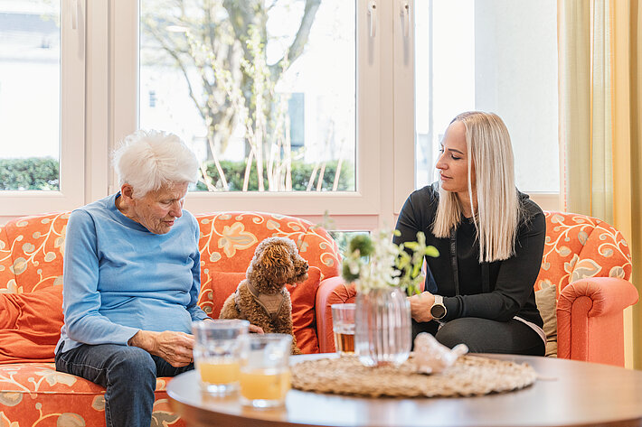 Ein ältere Frau und jüngere Frau sitzen auf einem orangefarbenen Sofa und schauen auf einen kleinen braunen Hund zwischen ihnen