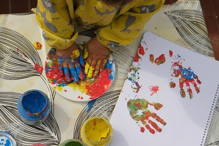 Ein Kind gestaltet mit bunten Fingerfarben Handabdrücke auf Papier.