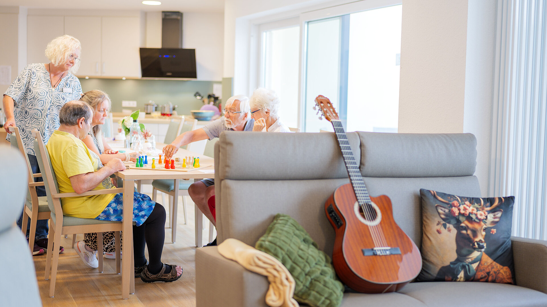 Seniorinnen und Senioren spielen gemeinsam am Esstisch einer Wohnküche, vorne ein Sofa mit Gitarre.