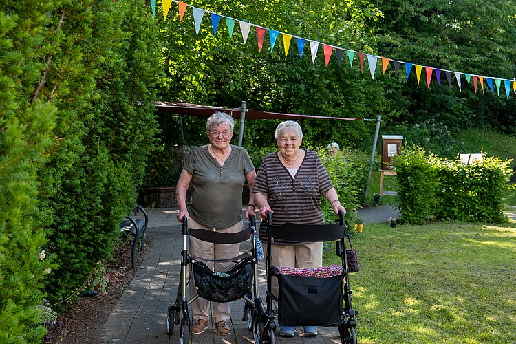 Zwei Seniorinnen gehen lächelnd mit ihren Rollatoren durch den Garten