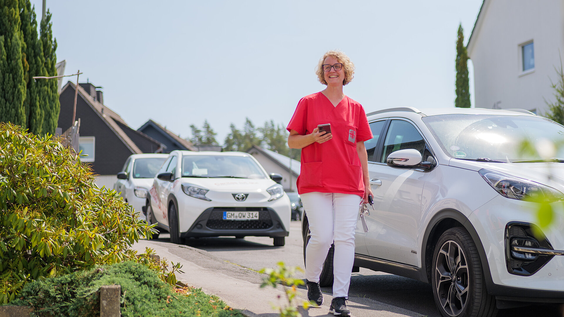 Pflegekraft der ambulanten Pflege geht lächelnd an Dienstwagen vorbei eine Wohnstraße entlang.