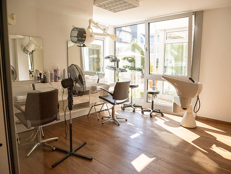 Heller Friseursalon mit zwei Stühlen, Spiegeln, einem Stehventilator und einem Waschbecken vor einem großen Fenster