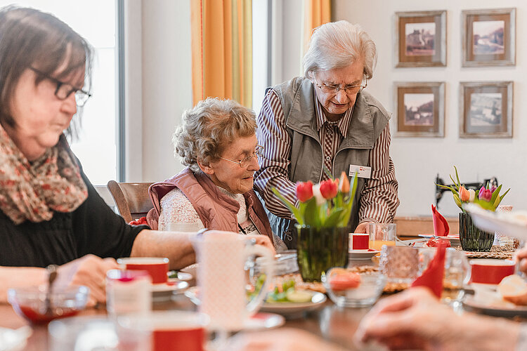 Eine Ältere Frau steht und spricht mit sitzender älterer Frau an einem gedeckten Tisch