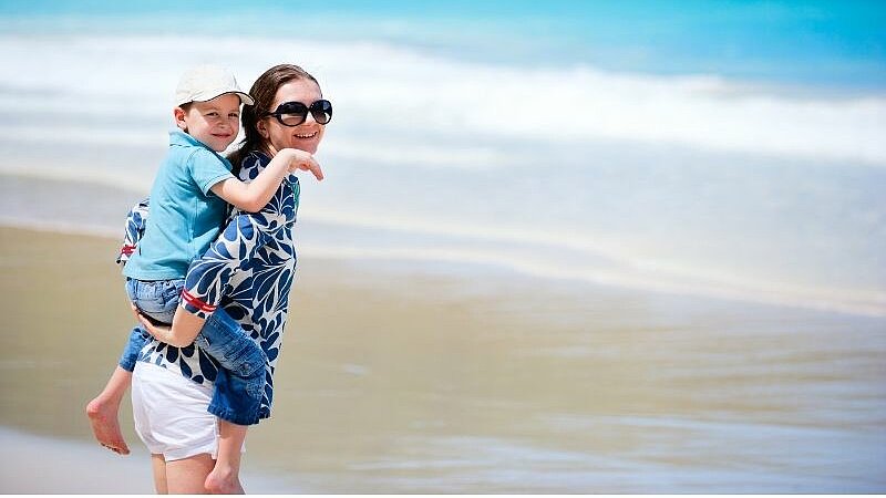 Eine erwachsene Person trägt ein Kind am Strand auf dem Rücken.