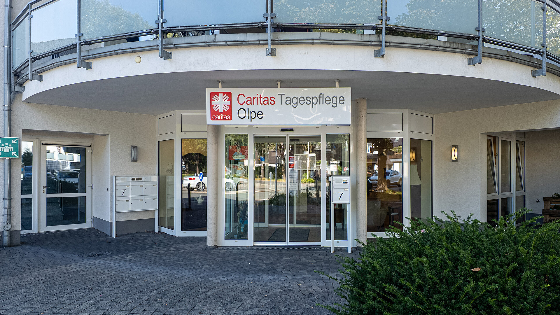 Eingangsbereich eines Gebäudes mit automatischen Glastüren und einem Schild mit der Aufschrift 'Caritas Tagespflege Olpe'