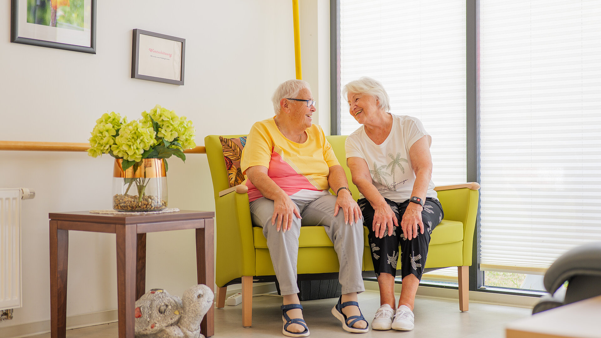 Zwei Seniorinnen sitzen lachend nebeneinander auf einem Sofa in einem hellen Aufenthaltsraum.
