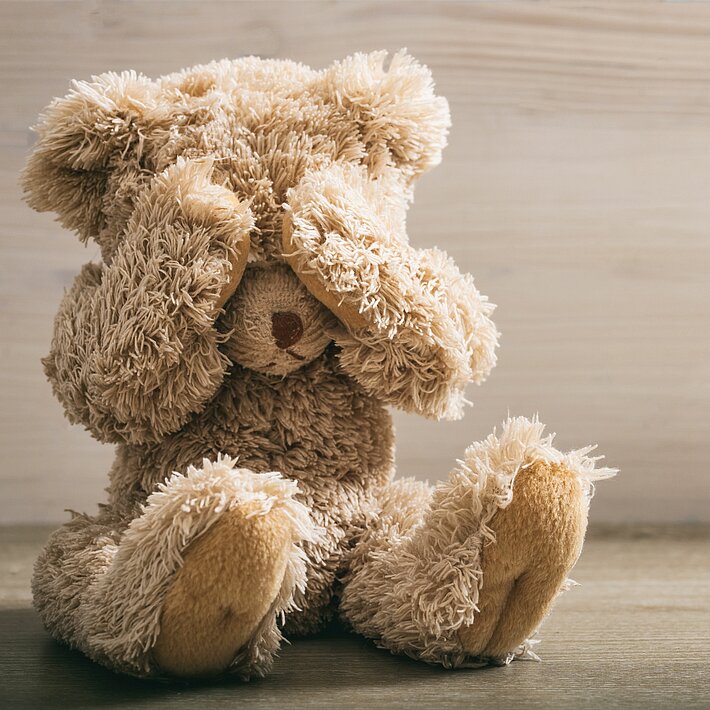 Ein brauner Teddybär sitzt allein auf dem Boden vor einer grauen Wand.