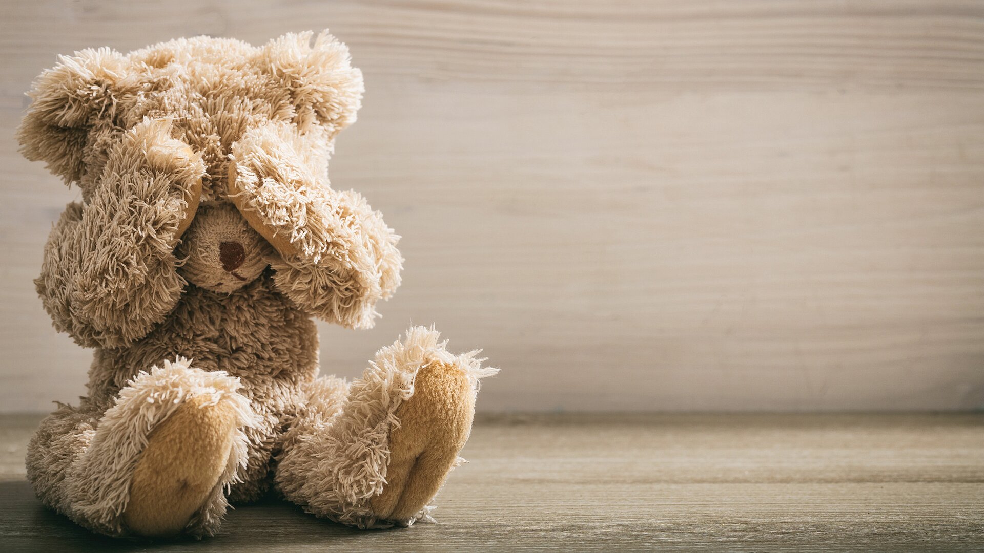 Ein brauner Teddybär sitzt allein auf dem Boden vor einer grauen Wand.