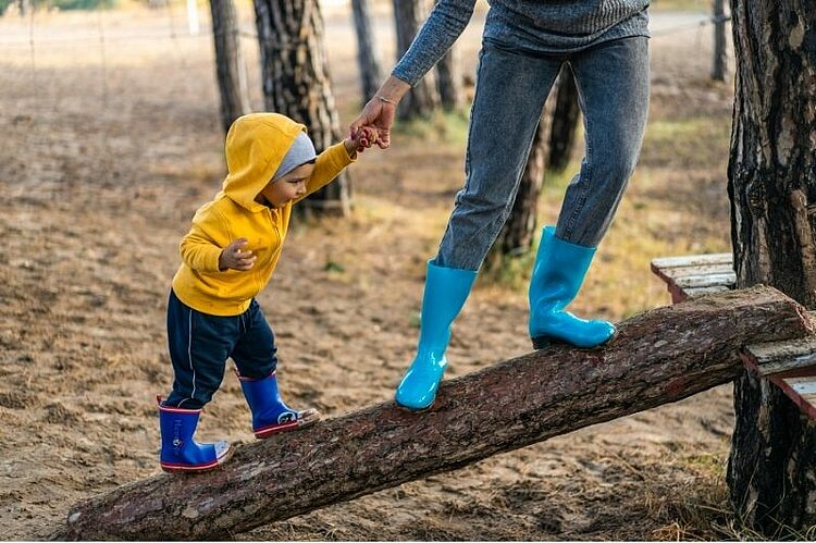 Eine erwachsene Person hält ein Kind beim Balancieren auf einem Baumstamm an der Hand.