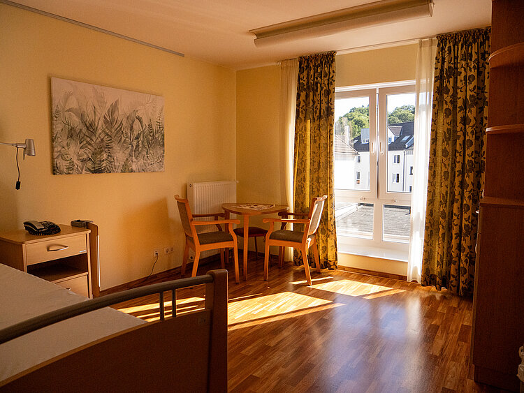 Ein Zimmer mit Holzboden, einem Bett, einem kleinem Tisch mit zwei Stühlen. Am Fenster sind gemusterte Vorhänge und an der Wand hängt ein Bild mit Pflanzenmotiv