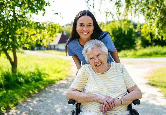 Eine junge Pflegerin schiebt eine Seniorin im Rollstuhl im Garten. Beide lachen.